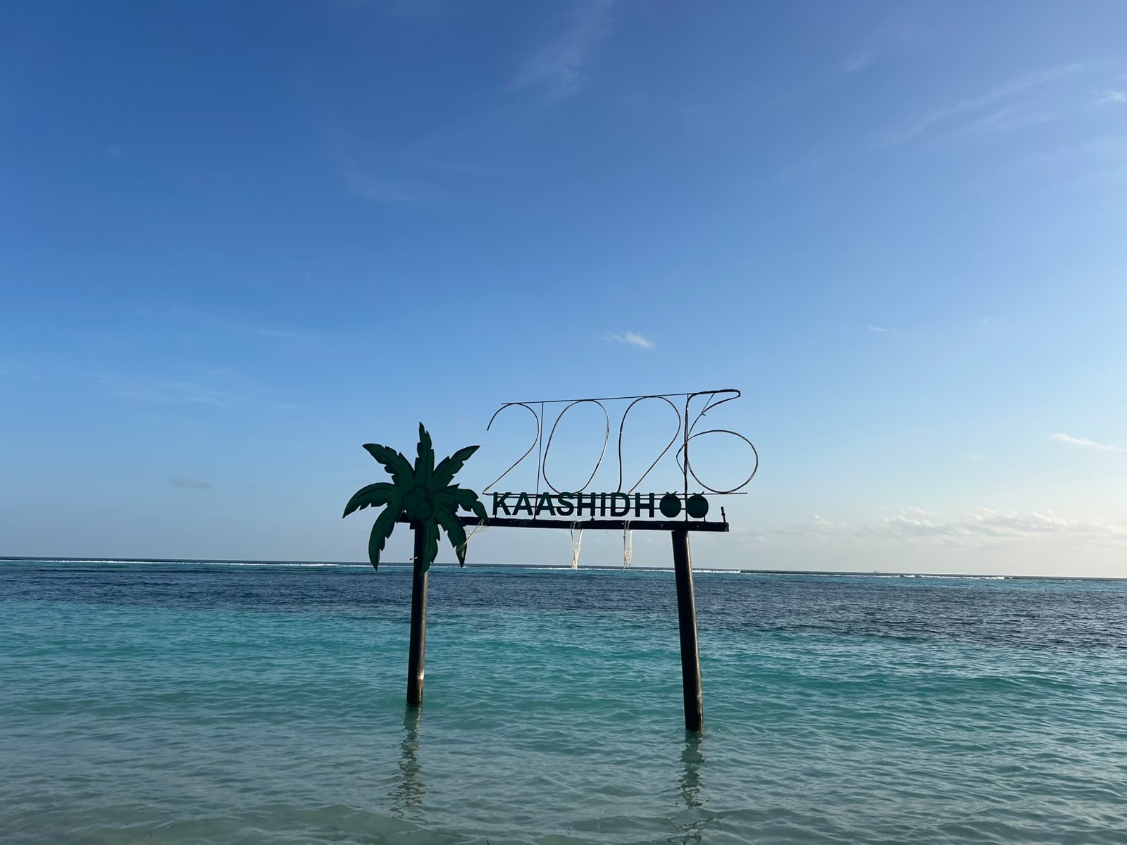 Metal 'KAASHIDHOO 2026' sign standing in shallow turquoise water off the reef, with an open sky overhead
